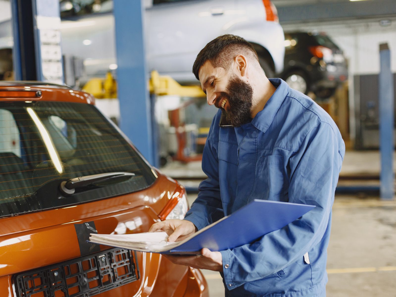 A smiling mechanic in coveralls inspecting a car and holding paperwork — illustrative stock photo, not a photo of Andy's own shop.