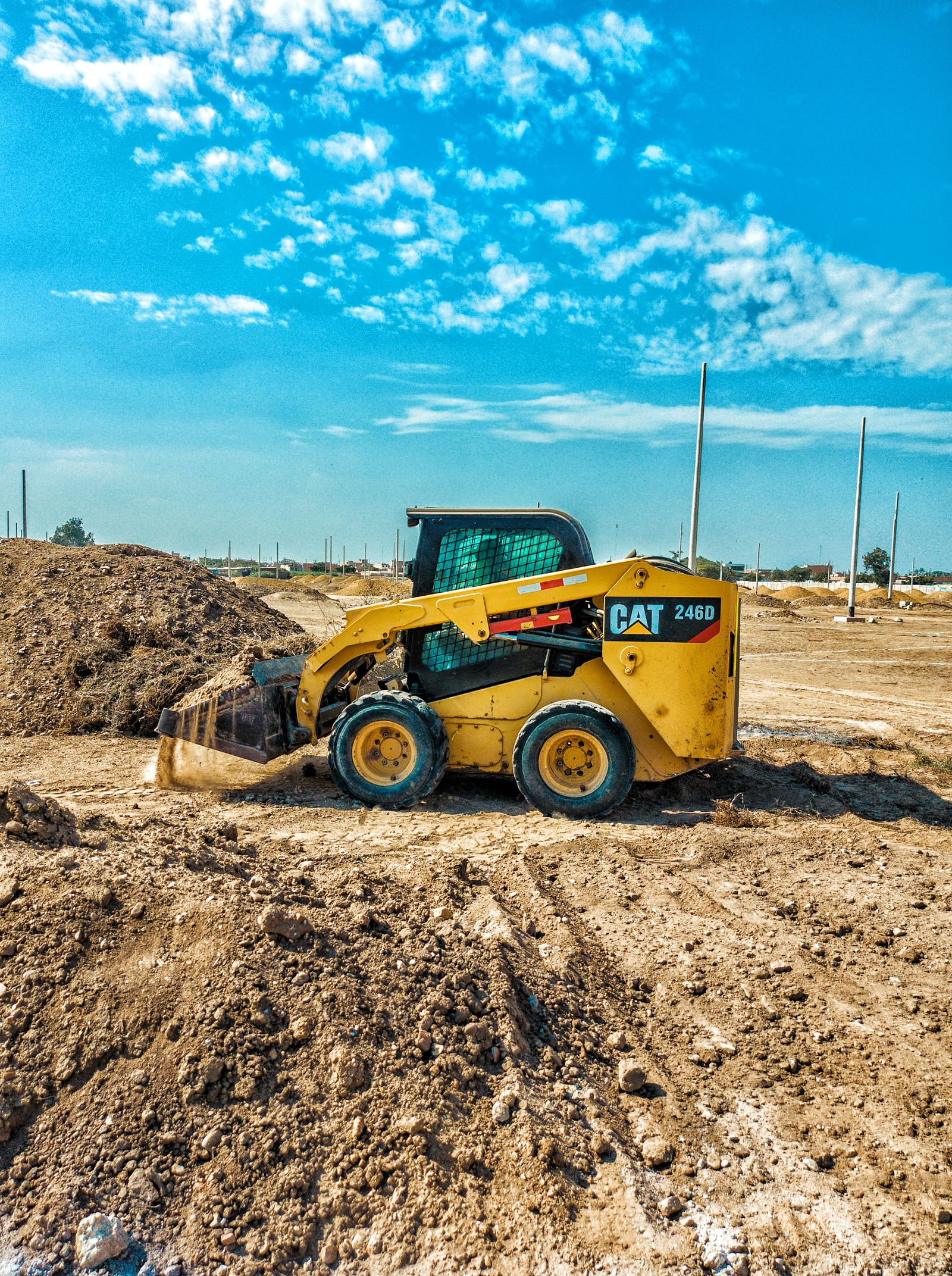 A yellow skid-steer loader moving brown earth on a construction site under a vivid blue sky with scattered clouds — illustrative stock photo, not Bernado's Landscaping and Dirtworks' own crew or equipment
