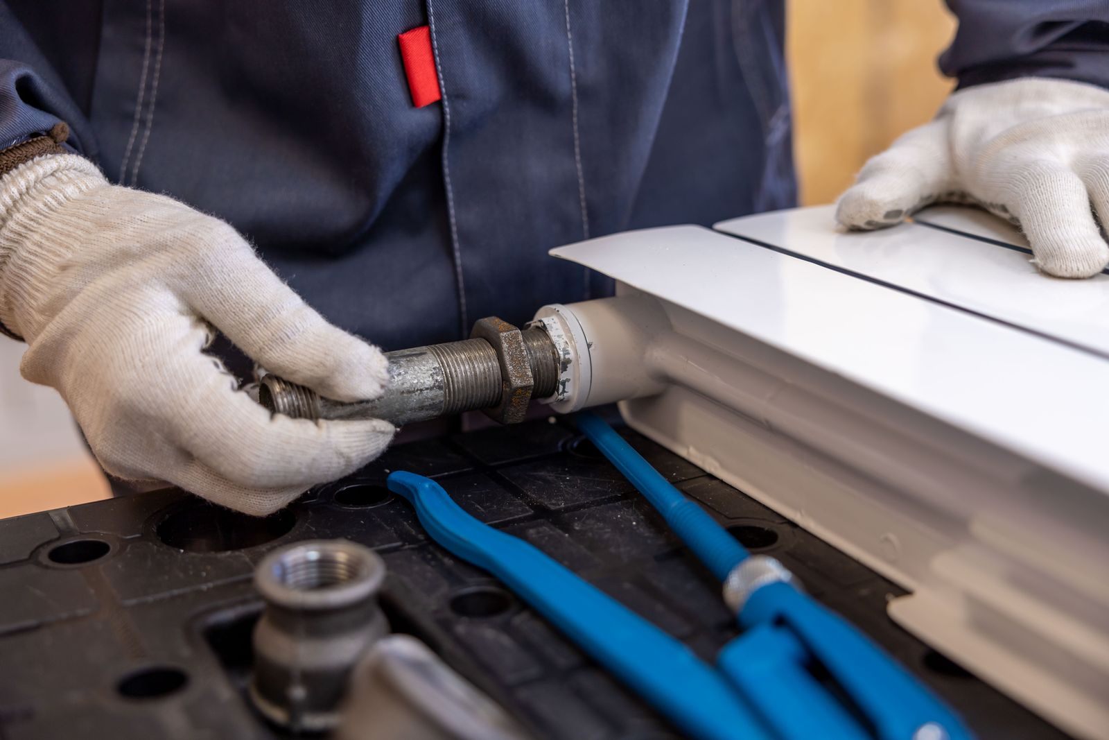 Gloved hands in a navy work uniform installing a pipe fitting onto a white radiator, blue-handled tools laid out on a dark work surface nearby &mdash; illustrative stock photo, not a photo of Dean Plumbing & Repair or its staff.