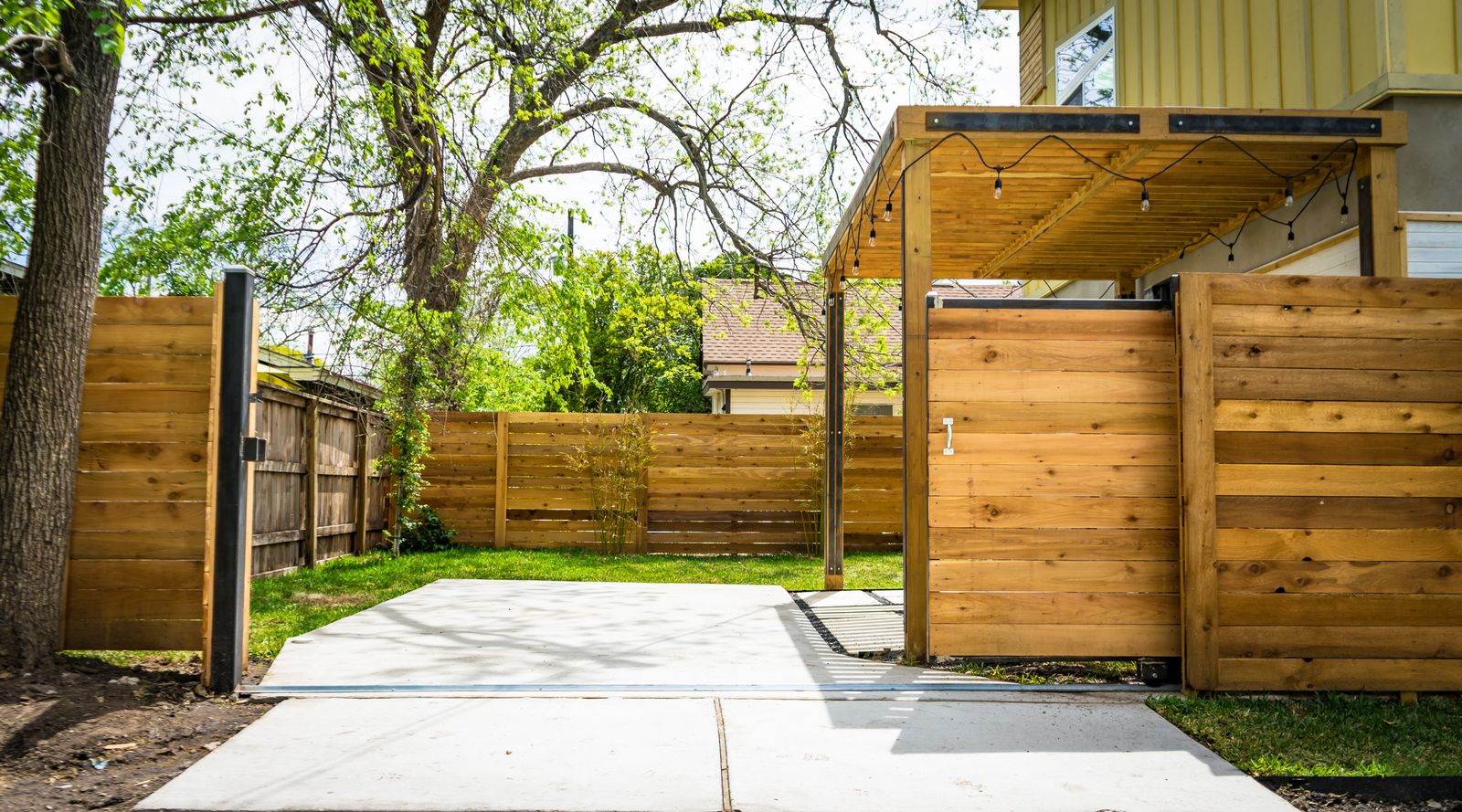 A modern horizontal cedar-plank privacy fence with a black metal gate frame, opening onto a clean concrete driveway — illustrative stock photo, not a photo of an MK Fencing job.